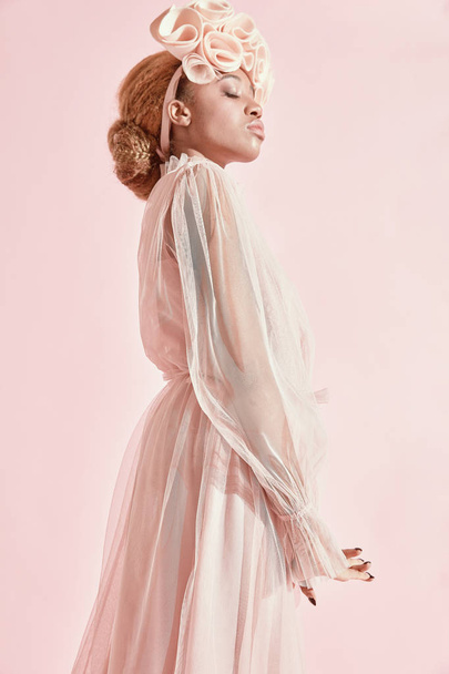 Studio portrait of black woman with light hair and green eyes wearing powder pink tulle dress and big frill hairband. Pink background - Photo, image