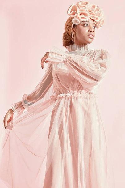 Studio portrait of amazing black woman with light hair and green eyes wearing powder pink tulle dress and big frill hairband and posing. Pink background - Photo, Image