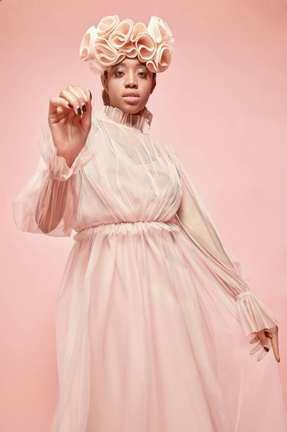 Soft studio portrait of beautiful black woman wearing powder pink tulle dress and big frill hairband and posing. Pink background - Foto, Imagen