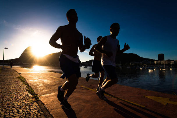 Siluetas de tres hombres corriendo en la madrugada durante la hermosa salida del sol caliente en Río de Janeiro con la montaña Sugarloaf en el horizonte
 - Foto, imagen