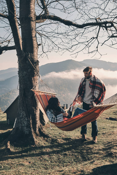 woman sitting in hammock in mountains, while handsome man bringing tea mugs - Photo, Image