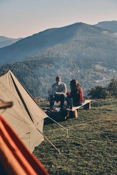 Caucasian couple in love sitting on bench near bonfire and tent in mountains with hot drinks in cups - Foto, afbeelding