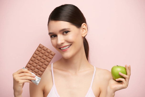 Smiling young pretty woman preferring bar of chocolate to green apple - Photo, Image