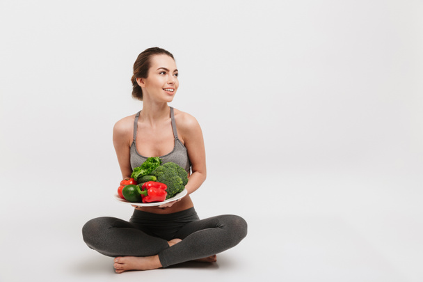 happy young woman sitting on floor with tray of various healthy vegetables isolated on white - Photo, Image