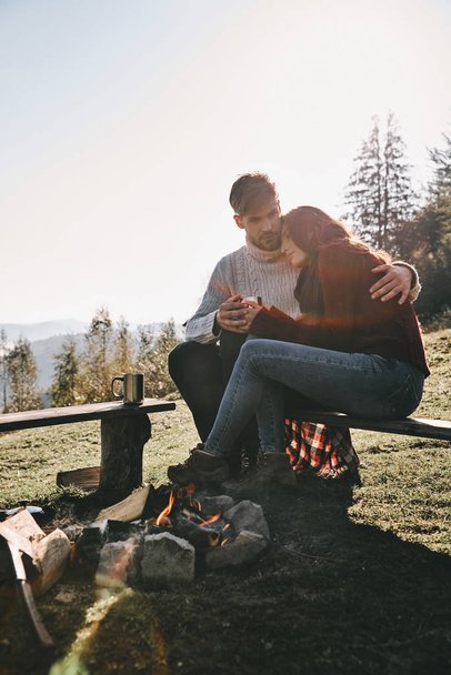 Caucasian couple in love sitting on bench near bonfire in mountains with hot drinks in cups - Foto, Imagen