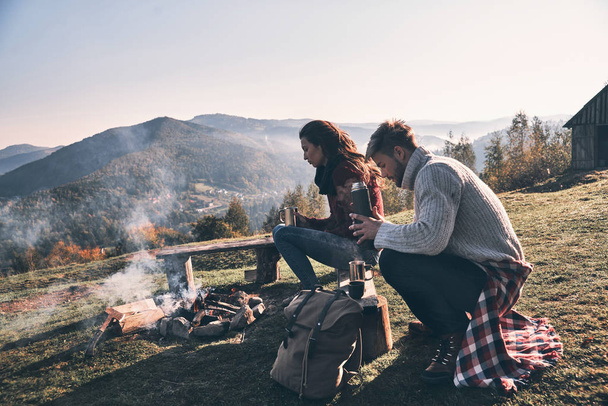 Caucasian couple in love sitting on bench near bonfire in mountains with hot drinks in cups - Foto, immagini