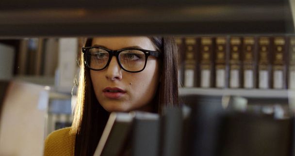 Bliska student młoda atrakcyjna kobieta w wybierając okulary książkę wśród książek na półkach w bibliotece. Portret strzał. Pomieszczeniu - Zdjęcie, obraz