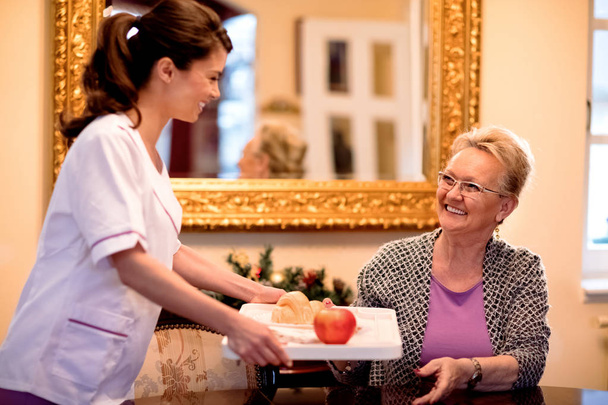 Smiling young nurse brings breakfast to senior woman at nursing  - Foto, Imagem