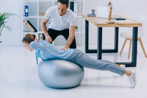 side view of businesswoman doing hyperextension exercise on fit ball with trainer at office - Fotografie, Obrázek