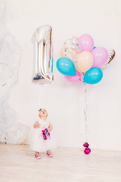 Sujeto cumpleaños de los niños es niños un año. Niña en habitación luminosa contra pared blanca en vestido y rubia sosteniendo bola inflable en forma de unidad, junto al grupo de bolas de diferentes colores
 - Foto, imagen