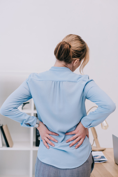 Rückansicht einer überlasteten Geschäftsfrau mit Rückenschmerzen, die sich im Büro auf einen Tisch lehnt - Foto, Bild