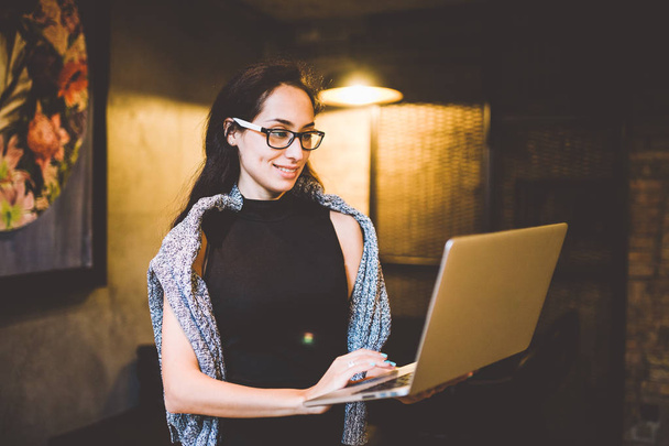 Het concept van kleine bedrijven en technologie. Jonge mooie brunette vrouw in zwarte jurk en grijze trui staat in Coffee Shop met laptop in handen. Maakt gebruik van glazen voor het zicht te typen op het toetsenbord - Foto, afbeelding
