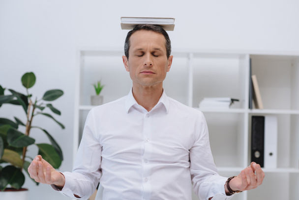 handsome businessman meditating with book on head at office - Photo, Image