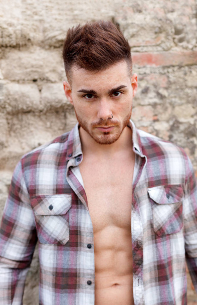 attractive guy wearing plaid shirt unbuttoned against wall of abandoned house - Photo, Image