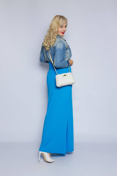 Studio portrait of a beautiful young blonde woman with curly hair. A sweet girl is dressed in a blue dress and a denim jacket, she is holding a white wicker bag in her hand - 写真・画像