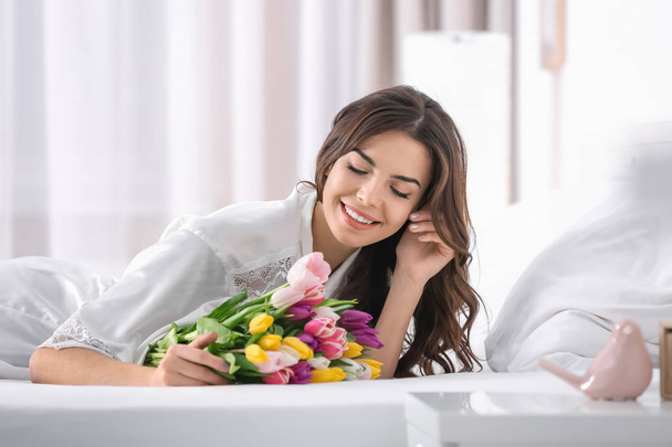 Beautiful woman with bouquet of tulips on bed at home - Foto, afbeelding