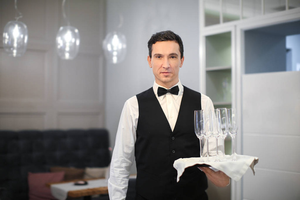 Молодой официант держит поднос со стаканами в помещении
 - Фото, изображение
