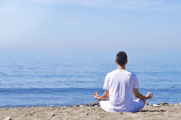 Молодой человек делает медитацию в позе лотоса на берегу моря / океана, гармонию и созерцание. Мальчик практикует йогу на морском курорте в отпуске. Стиль жизни. Мальчик отдыхает на открытом воздухе
. - Фото, изображение