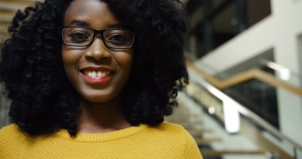 Close up van de krullend Afro-Amerikaanse mooie vrouw in glazen oprecht glimlachen naar de camera binnen modern gebouw. Portret - Video