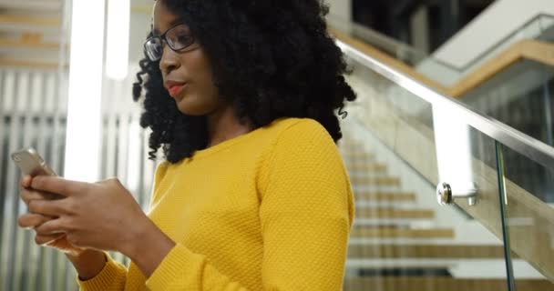 Young attractive African American curly woman in glasses smiling and chatting on the smart phone near big modern stairs. Close up. Portrait shot. Indoor - Záběry, video
