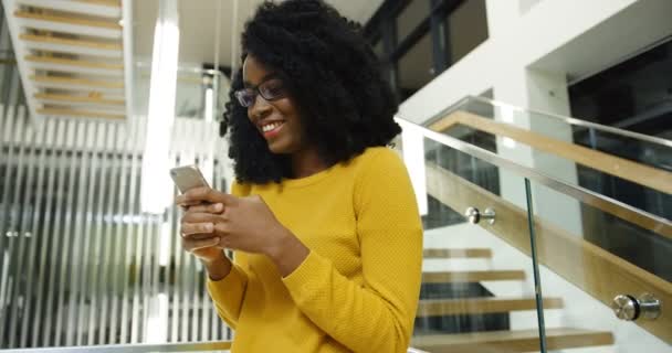 Young curly African American woman in glasses laughing while texting and getting messages on her smartphone near big urban stairs in the hall. Portrait shot. Indoors - Séquence, vidéo