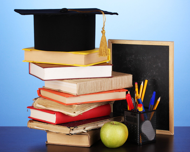 Books and magister cap against school board on wooden table on blue background - Photo, Image