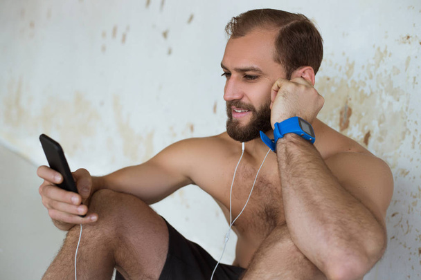 Όμορφος νεαρός χρησιμοποιώντας τηλέφωνο ενώ έχοντας άσκηση σπάσει στο γυμναστήριο. Ο τύπος χρησιμοποιώντας smartphone μετά την καθημερινή προπόνηση. Ο άνθρωπος που ακούς μουσική ενώ την ανάπαυση μετά την άσκηση. - Φωτογραφία, εικόνα