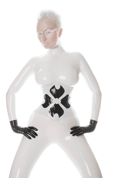 strange futuristic alien albino woman with short blonde hair posing in latex rubber catsuit and tight corset with black gloves on white background isolated - Photo, Image