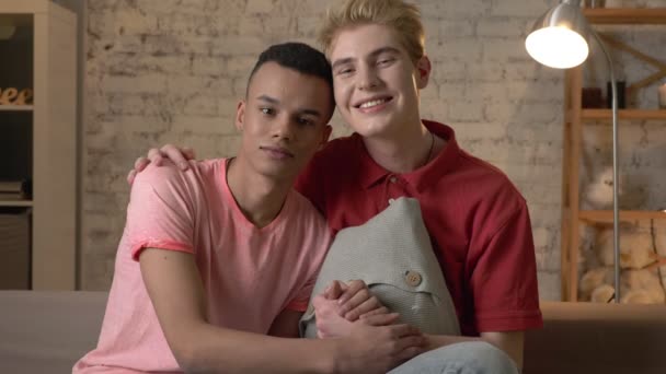 Dwóch wieloetnicznych homoseksualnych przyjaciół siedzieć na kanapie, trzymając się za ręce, przytulanie, uśmiechając się, patrząc na kamery. Przytulność domu, rodziny, lgbt. 60 fps - Materiał filmowy, wideo