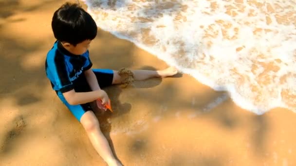 Chlapec v plavkách je posezení na pláži a hrál písek v moři v slunečný den v létě. - Záběry, video