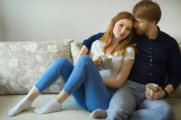 beautiful young couple guy and girl with red hair sitting on the couch and drinking tea - Fotoğraf, Görsel
