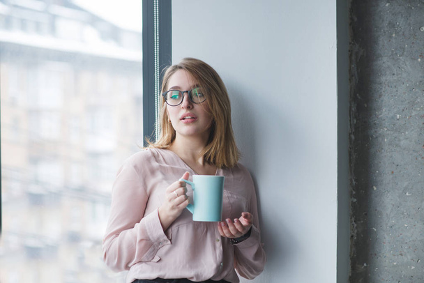 Красивая девушка в очках стоит в стене возле окна с чашкой кофе. Работница офиса девушки стоит с чашкой напитка в руках и смотрит в окно
 - Фото, изображение