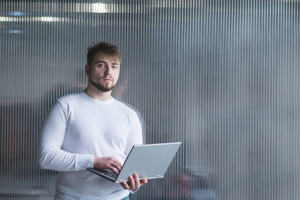 Un uomo è un impiegato in piedi sullo sfondo di un muro dell'ufficio con un computer portatile in mano. Uno sguardo alla telecamera
 - Foto, immagini