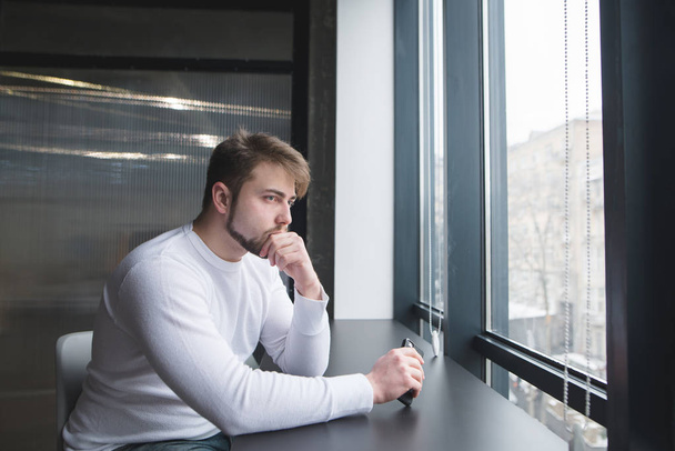 Un joven reflexivo se sienta en la oficina a la mesa y mira por la ventana. Un hombre piensa en la oficina en la mesa
. - Foto, Imagen