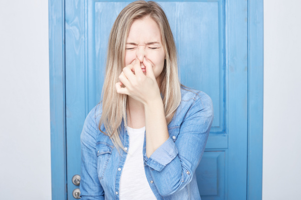Displeased European cute female student with blond hair female plugs nose as smells something stink, unpleasant, feels aversion, isolated over blue door background. Young woman hates disgusting scent. - Photo, Image