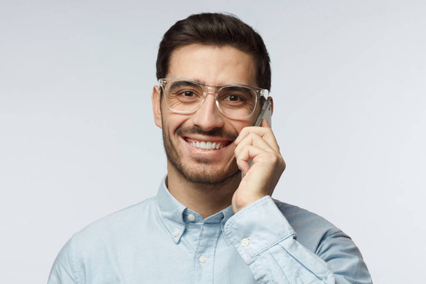Closeup shot of young man isolated on grey background wearing stylish transparent eyeglasses enjoying telephone conversation with happy toothy smile, laughing - Foto, Imagen