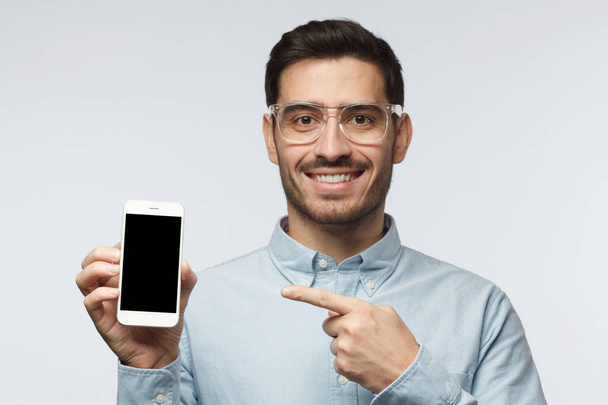 Shot of hansome man in blue shirt isolated on grey background, presenting smart phone and pointing with finger at blank black screen with copyspace for ads - Foto, imagen