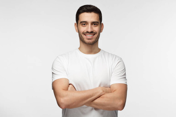 Portrait of smiling handsome man in white t-shirt standing with crossed arms isolated on gray background - Foto, imagen