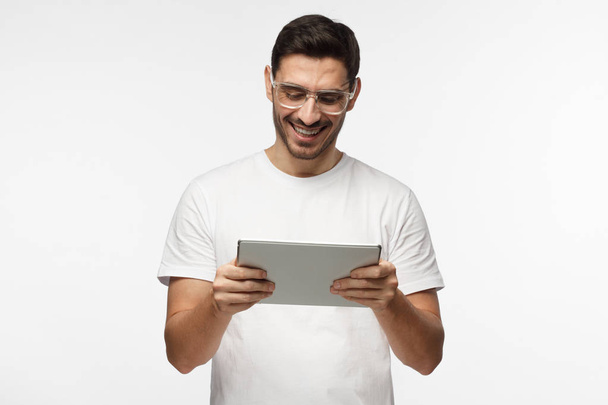 Studio closeup of young man pictured isolated on grey background wearing transparent eyeglasses looking attentively to screen of tablet he is holding, smiling happily, laughing - Фото, изображение