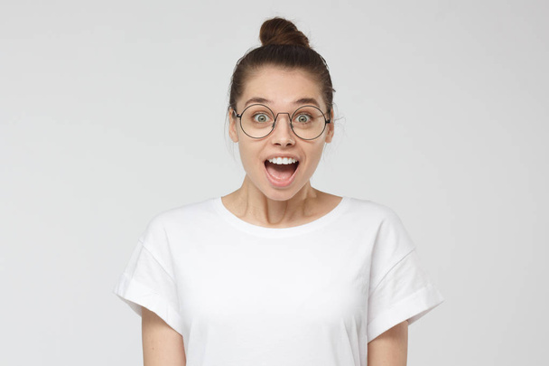 Indoor shot of attractive young woman with brown hair tied in bun isolated on grey background, smiling happily with mouth open in surprise, anticipating cool prospects in future with pleasure - Foto, Imagen