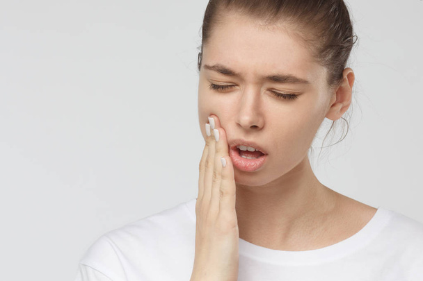 Young European woman isolated on grey background suffering from severe toothache, feeling pain so strong that she is pressing fingers to cheek to calm it down, looking desperate - Photo, Image