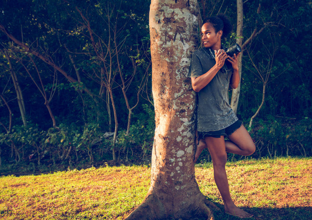 Afro americký mladá dívka schovává s dslr fotoaparát za starý velký strom v parku - Fotografie, Obrázek