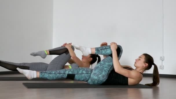 In the gym during the pilates exercise lying on the gym mats two girls lying on their back alternately pull their knees to the chest stretching the muscles of the thighs. Synchronous execution of the - Кадри, відео