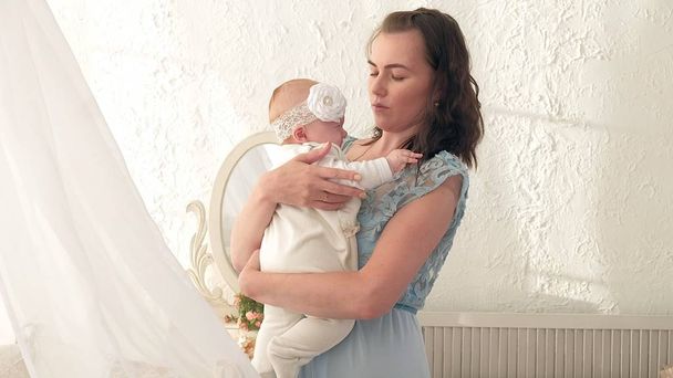 Seguindo a mãe colocando o bebê para dormir enquanto caminha. Mulher atraente segurando bebê em mãos e andar através da sala de estar com cortinas de janela brilhantes no fundo
. - Foto, Imagem