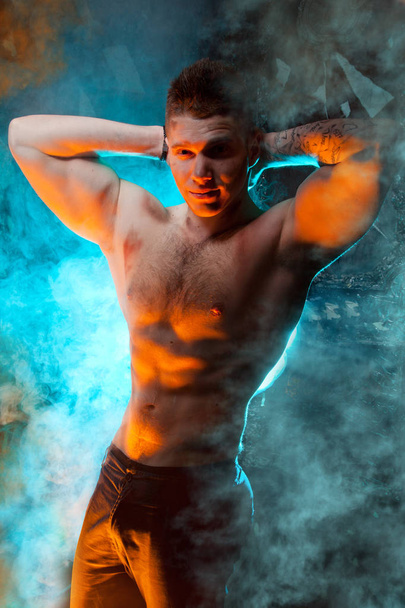 чоловічий молодий м'язистий чоловік стоїть з голим торсом у диму на чорному тлі в студії
 - Фото, зображення