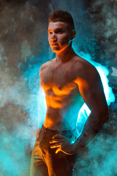 masculine young muscular male standing with naked torso in the smoke on a black background in Studio - Fotó, kép