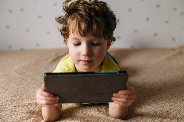 Little cute boy in a green T-shirt playing games on a tablet and watching cartoons. Toddler with tablet. - Fotografie, Obrázek