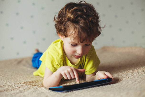 Little cute boy in a green T-shirt playing games on a tablet and watching cartoons. Toddler with tablet. - Fotografie, Obrázek