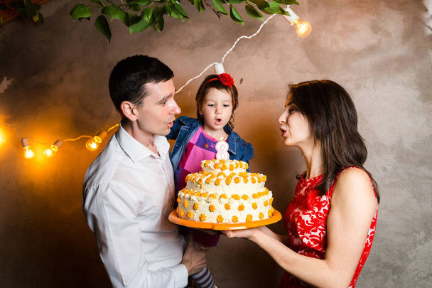 Thema Familienurlaub Kindergeburtstag und Ausblasen von Kerzen auf großen Kuchen. junge dreiköpfige Familie steht und hält 5-jährige Tochter im Hof gegen graue Wand und Girlanden gelbe Lampen - Foto, Bild