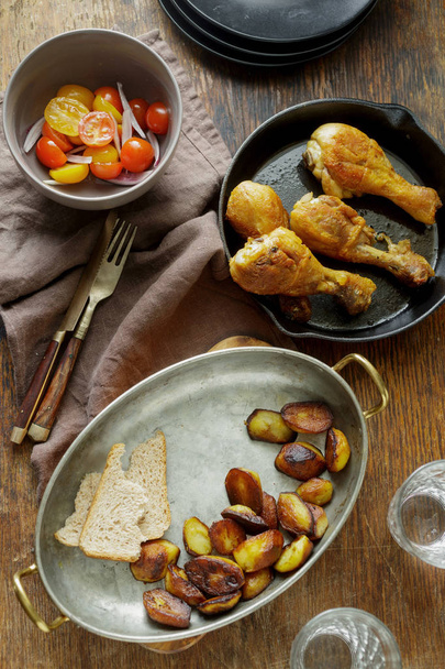 Fried chicken legs in frying pan on dark wooden table with potatoes and salad. Dinner table concept - Photo, image